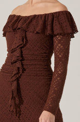 A woman is wearing an elegant off-the-shoulder dress made of brown lace fabric. The dress features a large ruffle that runs along the neckline and extends around the shoulders, adding a decorative touch. The long sleeves are fitted and made from the same lace material, which has a floral pattern. The dress is form-fitting, with ruching details on the sides.
